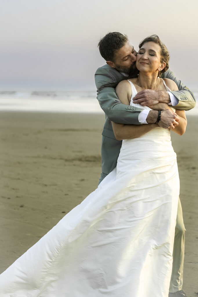 wedding couple wrapped up in a big hug with woman's back to man's chest. He is kissing her on the cheek