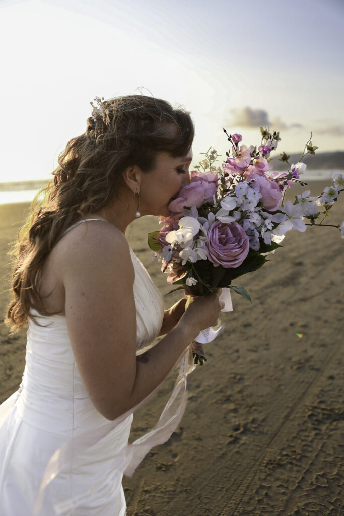 bride on beach smelling her bouquet of pink and white flowers. 