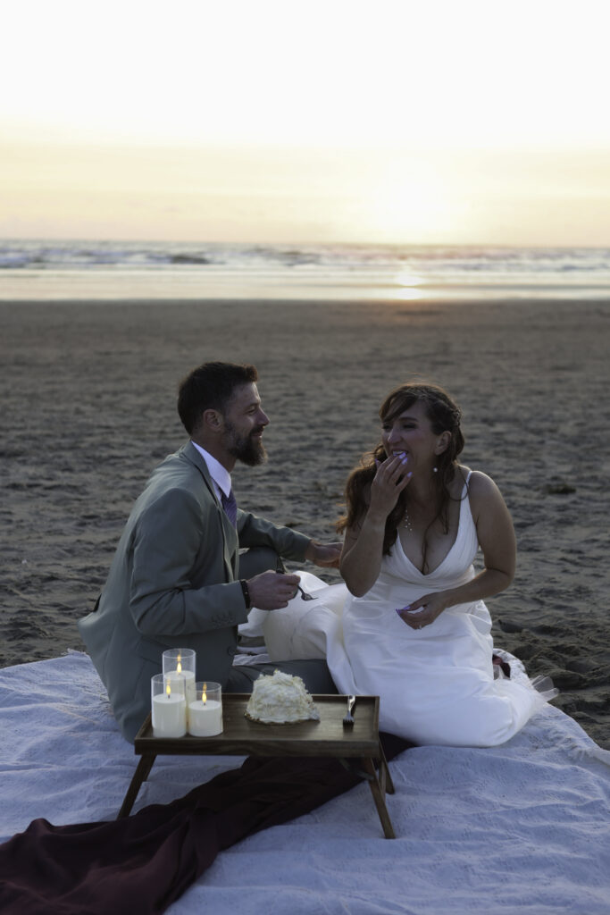 wedding couple has picnic on the beach. they laugh as he feeds her cake