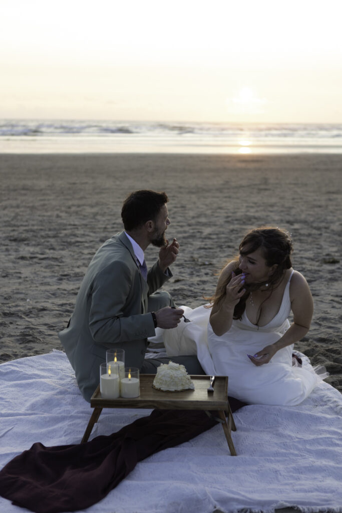 wedding couple has picnic on the beach. they laugh as he feeds her cake