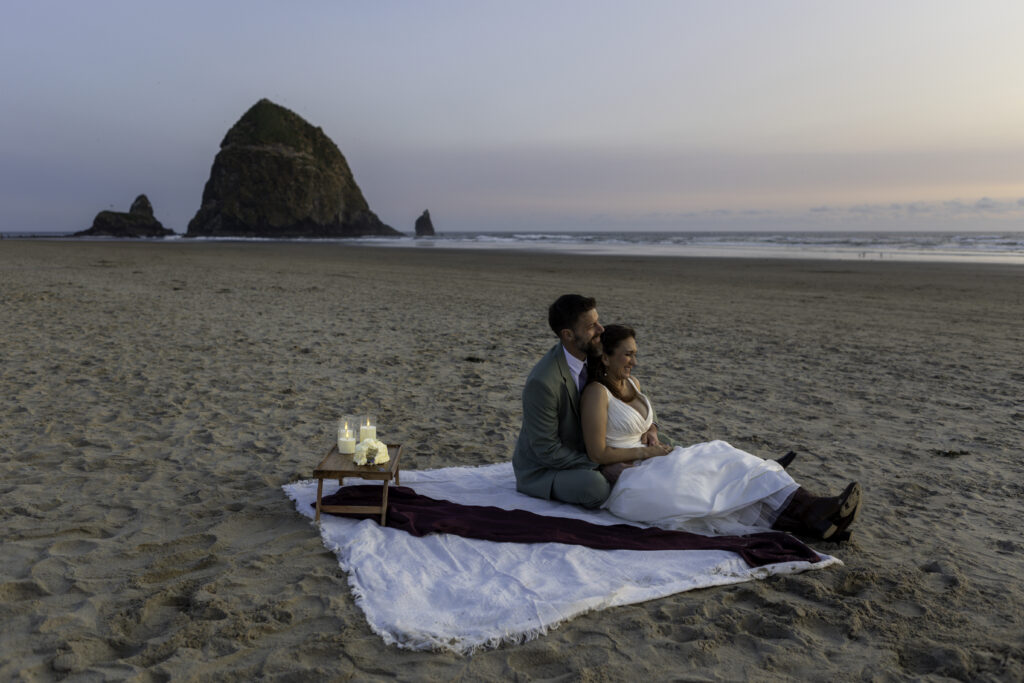 wedding couple has picnic on the beach. They are sitting with man in back and woman sitting in front of him. they are facing the ocean and enjoying the surf and sunset. 