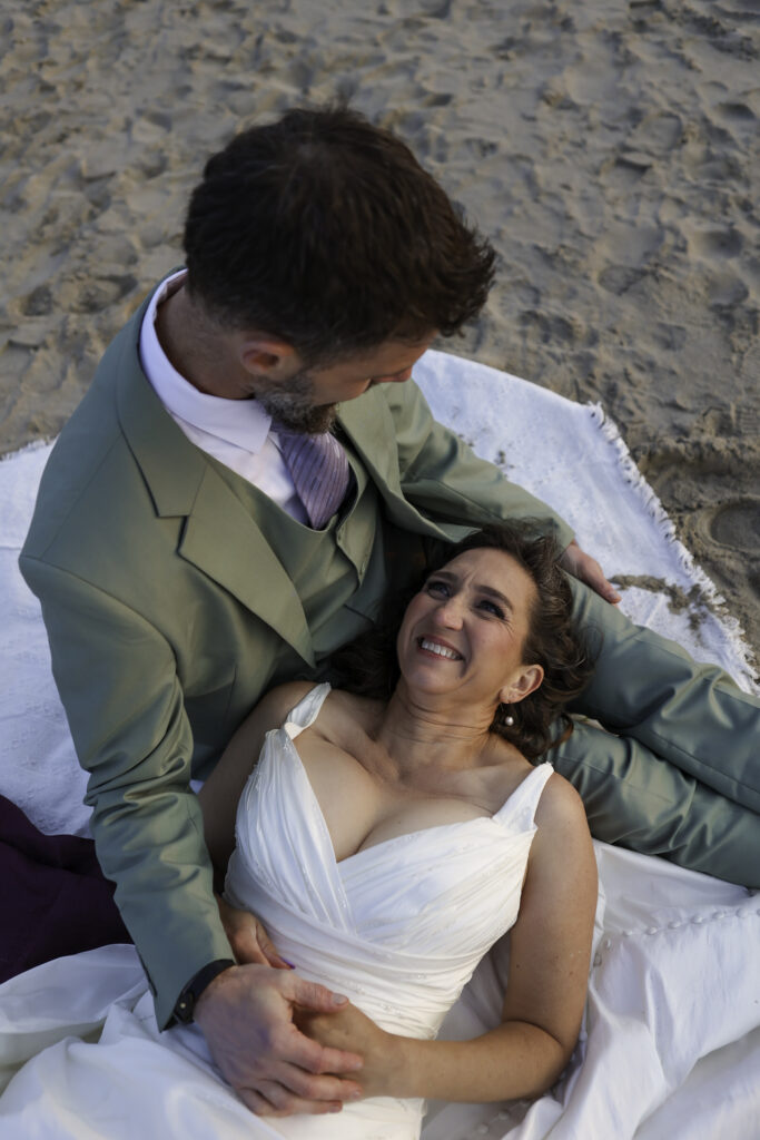 wedding couple has picnic on the beach.he sits  and she is laying with her head in his lap. they are looking at each other lovingly. 