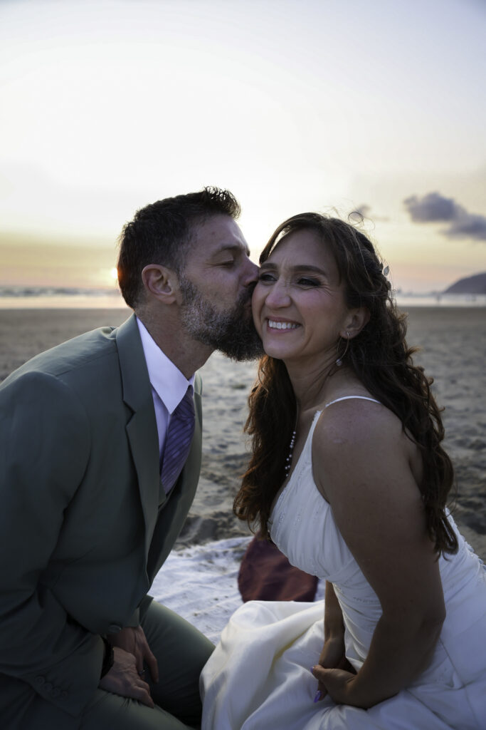 wedding couple has picnic on the beach. woman is sitting with her hands on her lap. Husband leans over to kiss her on the cheek.