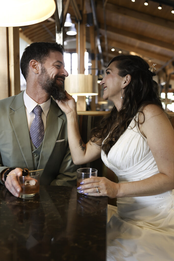 couple sitting at bar with cocktails with bride's hand under grooms chin. both smiling at each other 