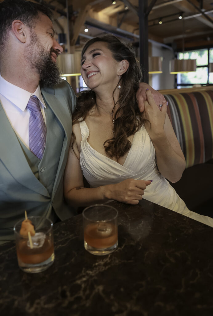 couple sitting in bar booth in wedding attire enjoying cocktails. Husband has arm wrapped around wife's shoulder and they look at each other lovingly