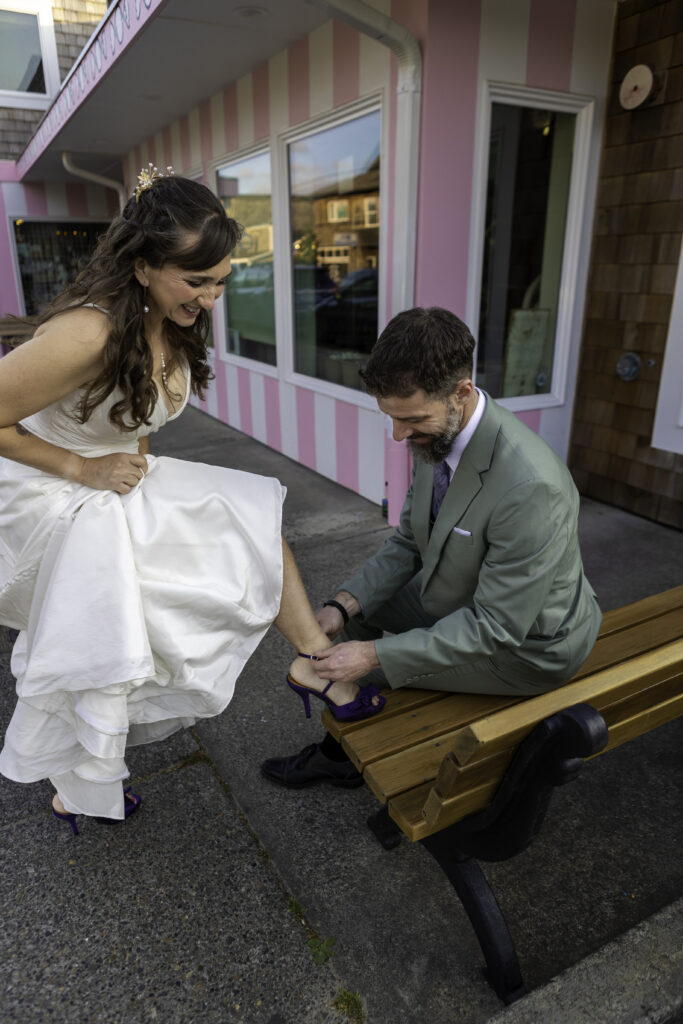 husband helps his wife secure her purple high heels while sitting on a bench. 