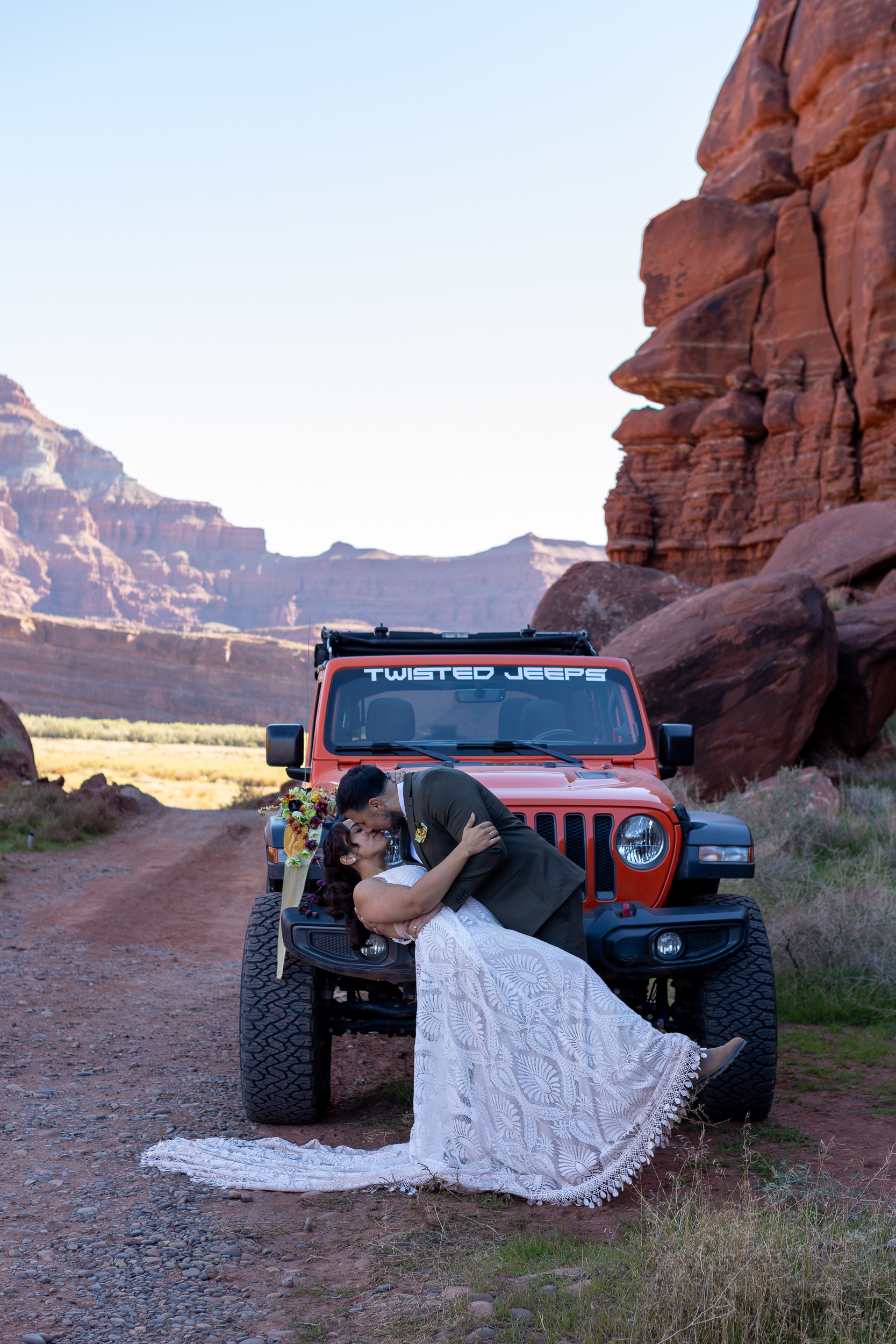 couple does a dip kiss in front of their orange jeep on Moab desert road. 
