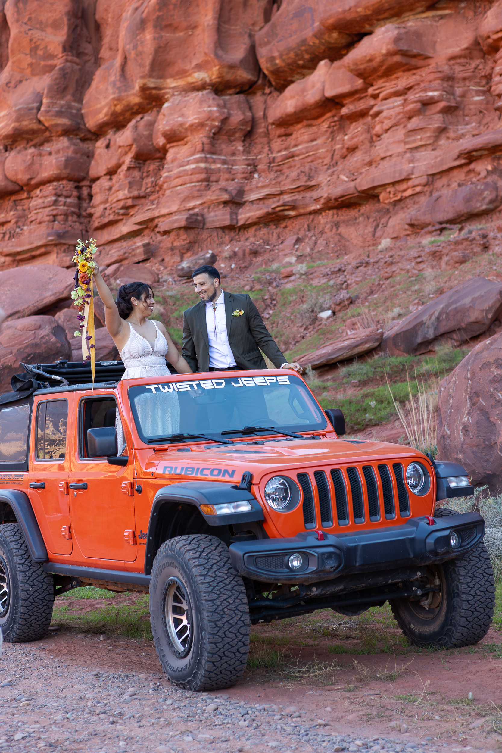 they have a moment of celebration as laura lifts her bouquet in the air as they look at each other while standing in their rental jeep