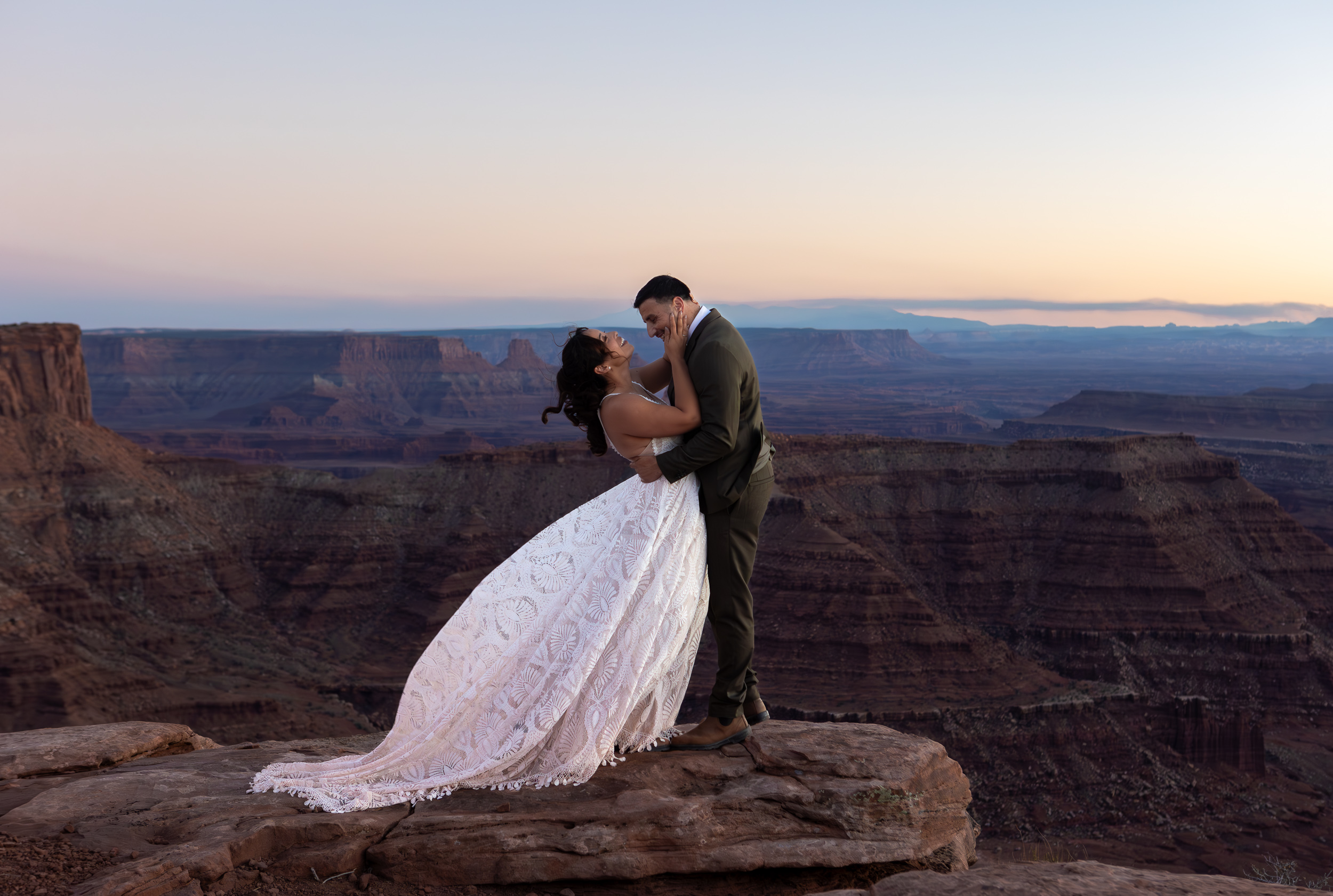 bride and groom stand facing each other with moab landscape in background. dress is flying in the wind. she has her back arch and her head thrown back in a big laugh.