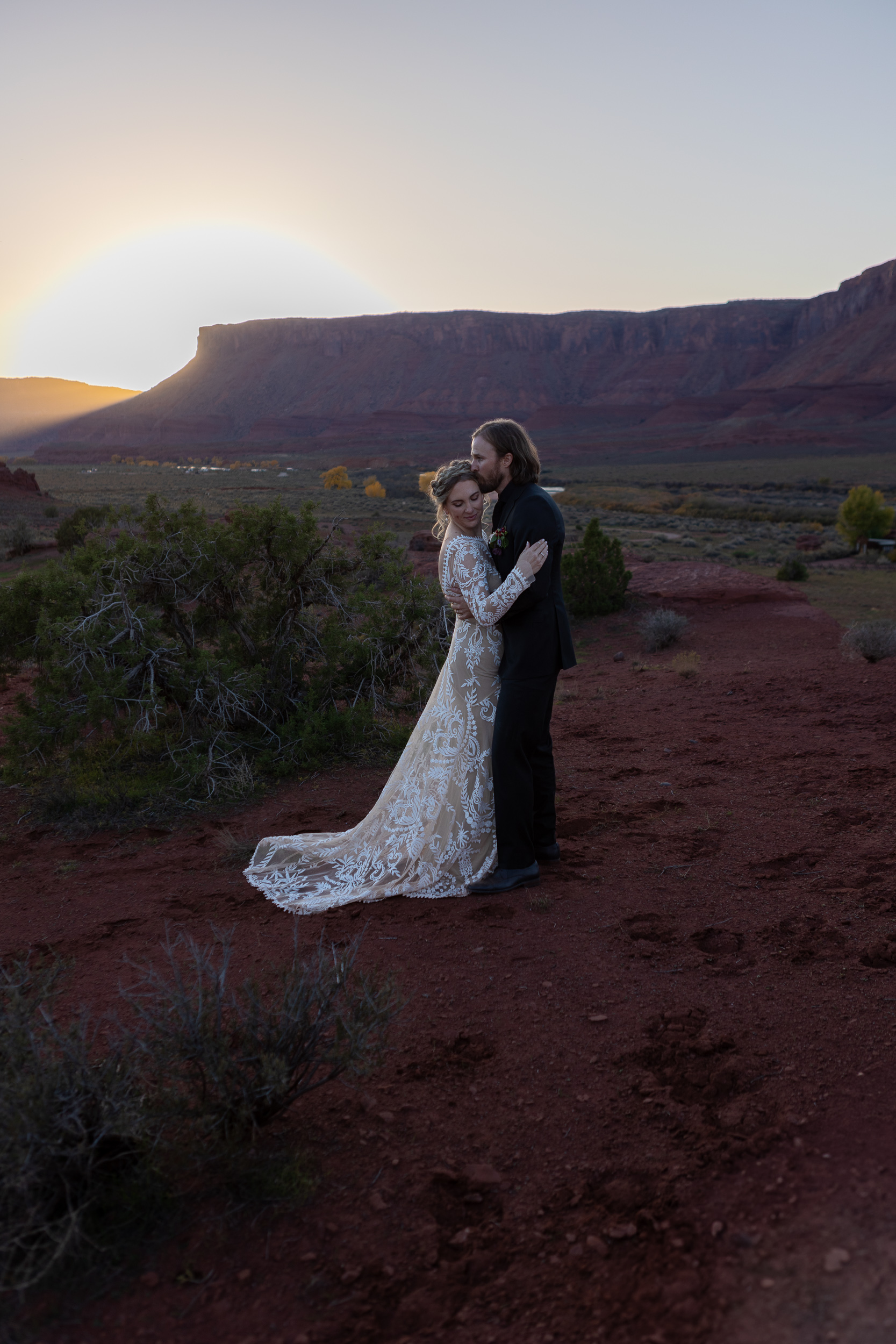 couple holding each other and husband kisses her temple with sun setting behind mountains in moab