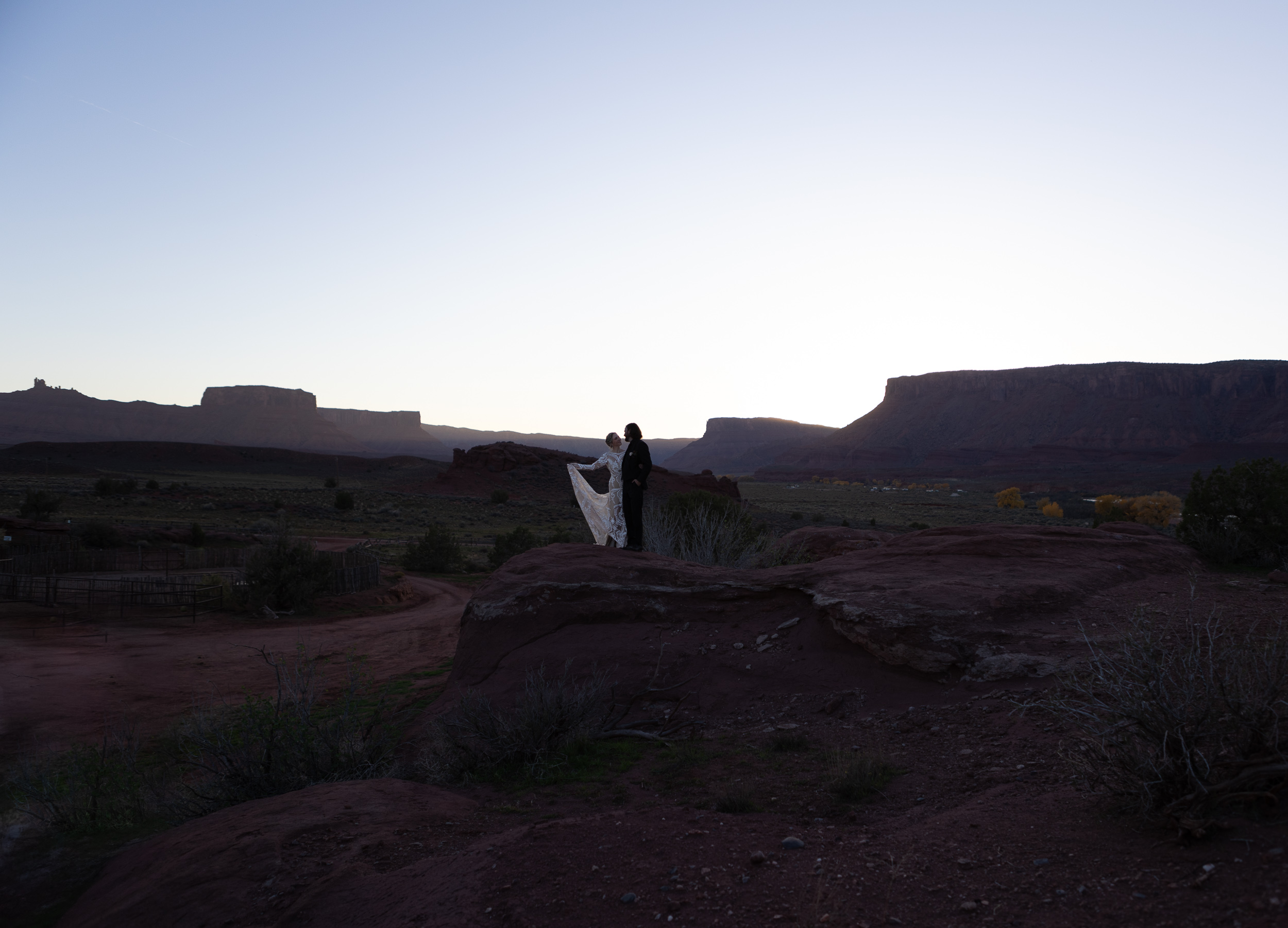 couple pose on rock with sun setting behind mountains in moab with bride holding dress up on right side