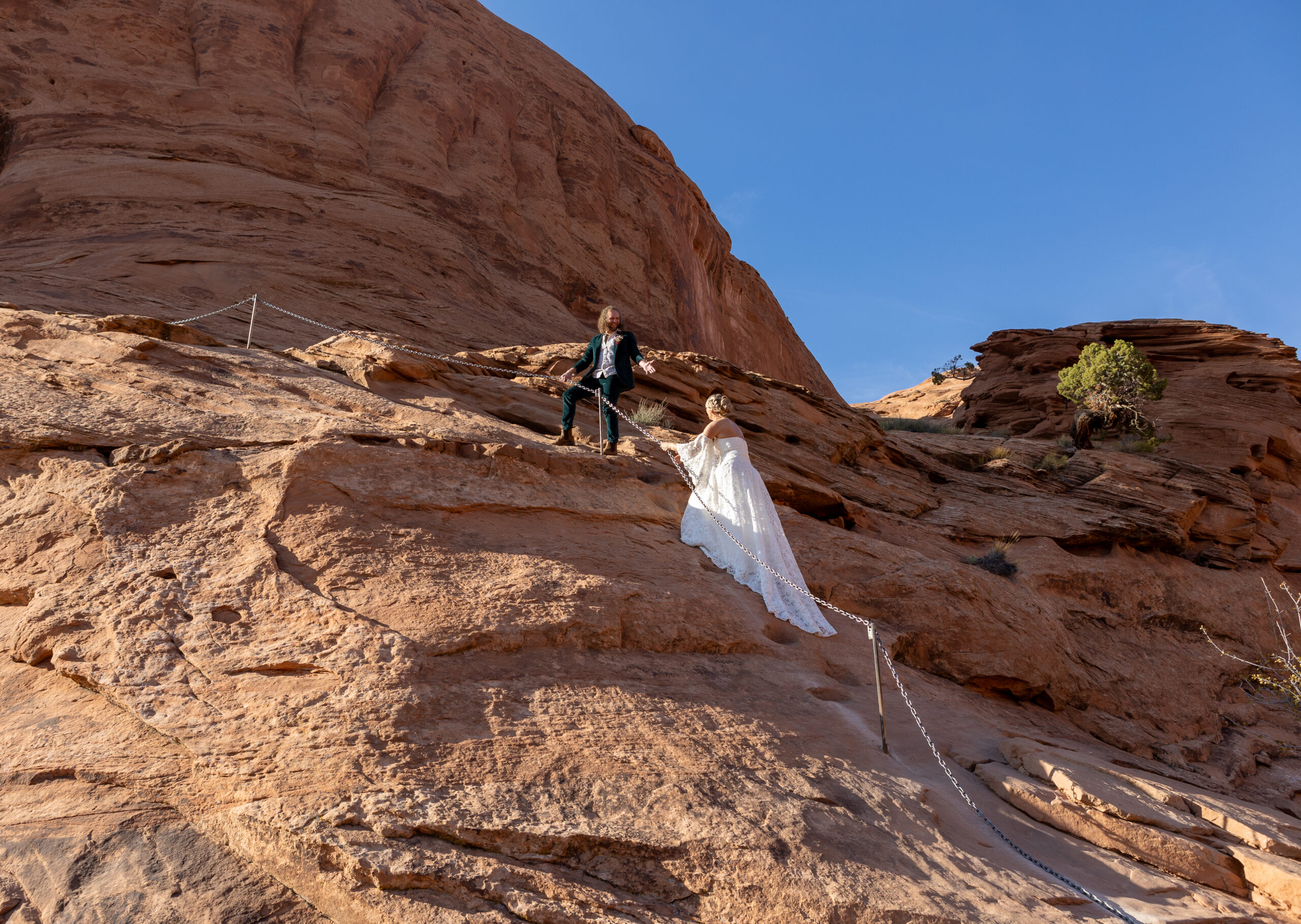 groom holding his hand out to help his bride up a cliff while they hike in Moab