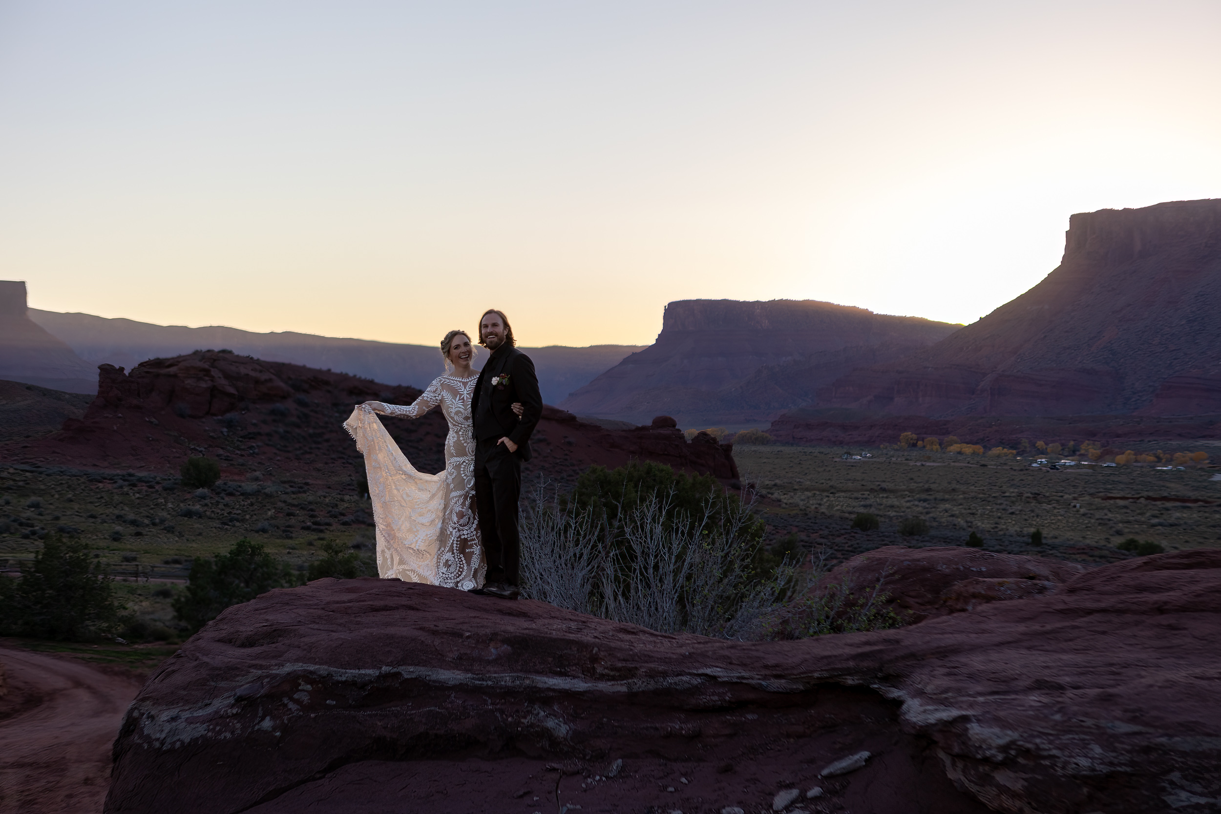 couple pose on rock with sun setting behind mountains in moab