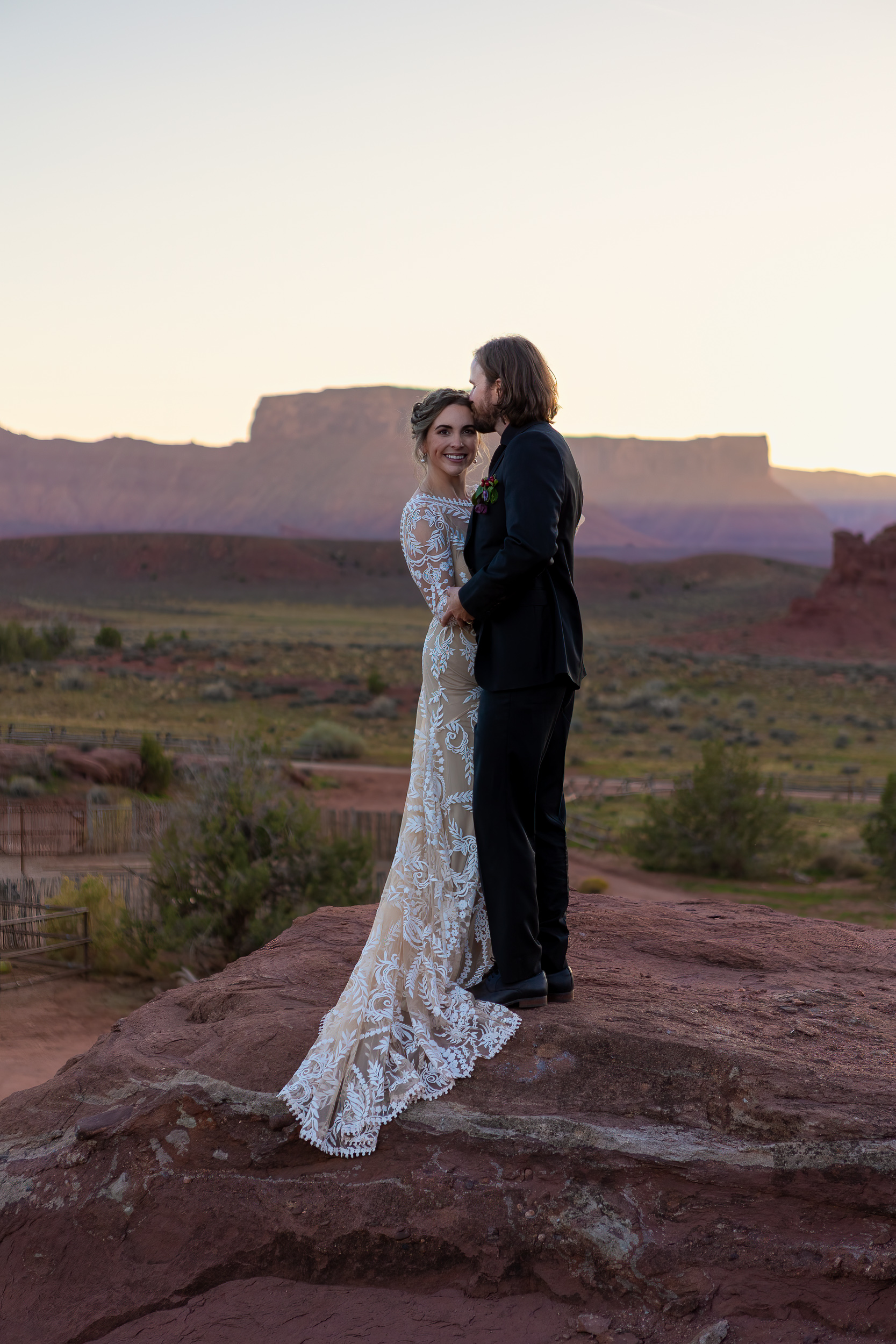 couple pose on rock with sun setting behind mountains in moab while husband kisses wifes temple