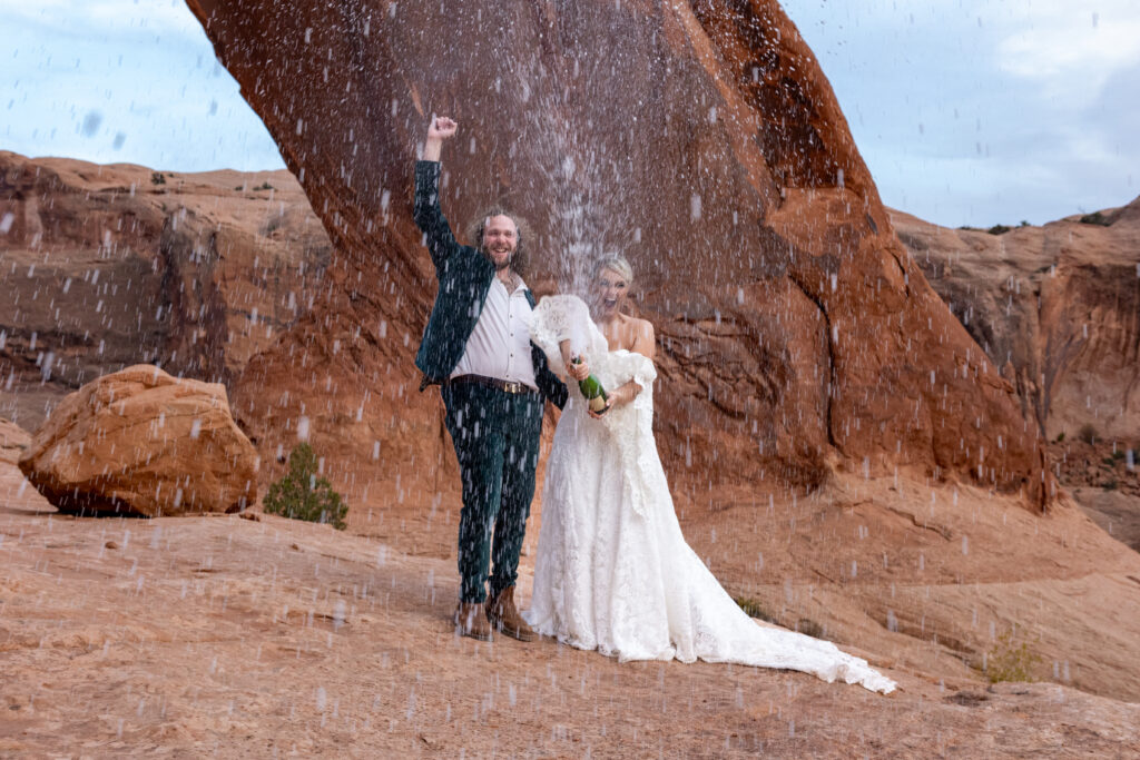 bridal couple celebrating their elopement with a champagne pop as bubbles fly in the air and groom pumps fist in the air