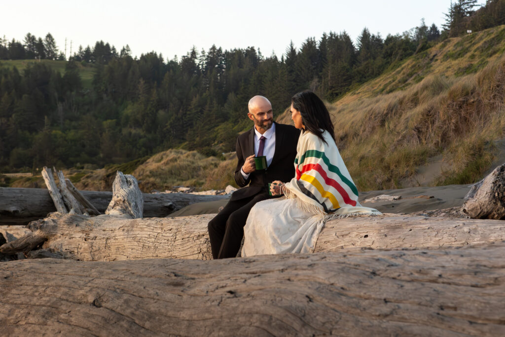 bridal couple sitting on a log in Brookings Oregon enjoying the sunset and sipping on hot chocolate as the sun sets.